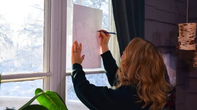 En kvinna som står med ryggen mot kameran och håller ett vitt papper mot ett fönster för att kunna teckna av ett mönster med hjälp av ljuset utifrån.