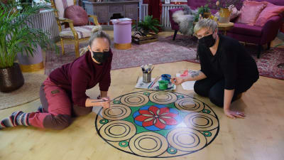 Två blonda kvinnor med ansiktsmasker som sitter på golvet i ett rum och målar en stor mandala på laminatgolvet.