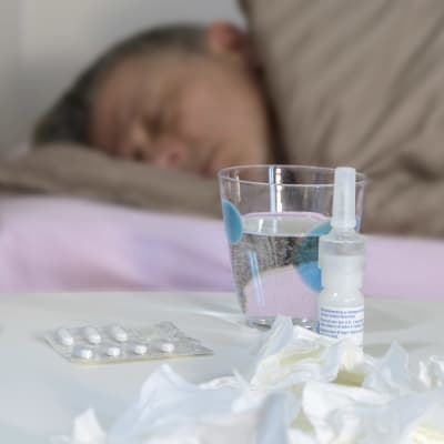 I förgrunden finns mediciner och näsdukar på ett bord, i bakgrunden ligger en förkyld man i sin säng