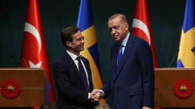 Sveriges statsminister Ulf Kristersson skakar hand med Turkiets president Recep Tayyip Erdoğan. Bakom de två männen syns en rad med flaggor, varannan turkisk och varannan svensk.