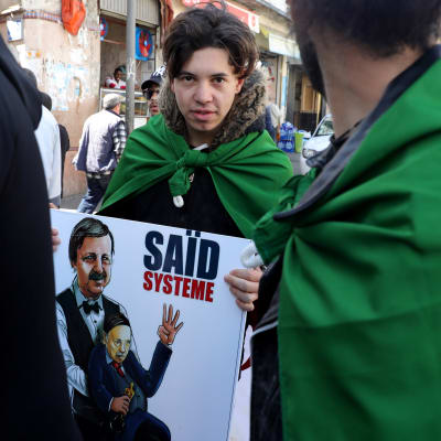 En man står med ett plakat i handen där Saïd Bouteflika ser ut att leda algeriets forna president