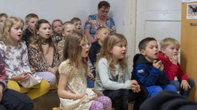 Förskolebarn sitter på bänkar i ett rum och sjunger tillsammans med en vuxen.