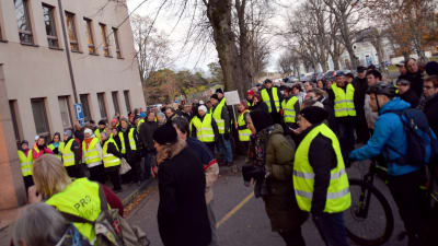 En stor grupp människor i gula reflexvästar står och demonstrerar framför en byggnad.
