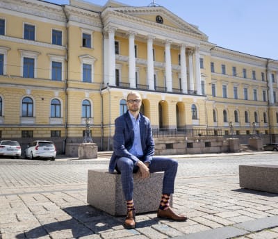 Mika Niemelä sitter på en sten på ett torg. I bakgrunden Finansministeriets byggnad.