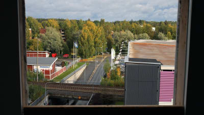 Rakennuksen ikkunasta avautuu näkymä, jossa rautatie ylittää tien. Vasemmalla teollisia rakennuksi, oikealla parkkihalli. Taustalla metsää. 