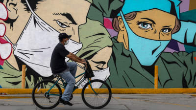 En person med ansiktsskydd på cyklar. I bakgrunden syns en vägg som är täckt med ett stort konstverk av personer med ansiktsskydd på.