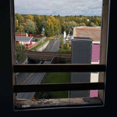 Rakennuksen ikkunasta avautuu näkymä, jossa rautatie ylittää tien. Vasemmalla teollisia rakennuksi, oikealla parkkihalli. Taustalla metsää. 