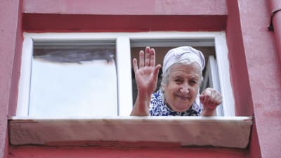 Glad äldre kvinna tittar ut genom fönstret och vinkar. 