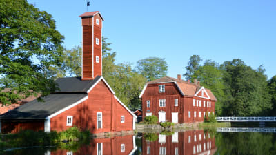 Två röda träbyggnader vid vatten, somrigt.