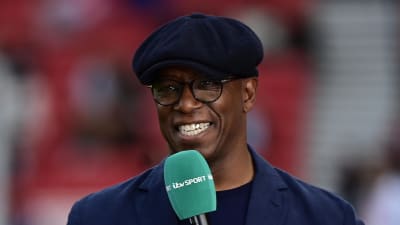 A smiling Ian Wright holds a green microphone.  He wears a blue jacket and hat.
