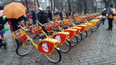 Ett 50-tal lånecyklar är parkerade på Domkyrkotorget i Åbo. Folk står med paraplyer och väntar på att få börja använda dem.