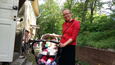 ung mamma med röd blus står med baby i barnvagn utanför ett trähus vid en grönskande park