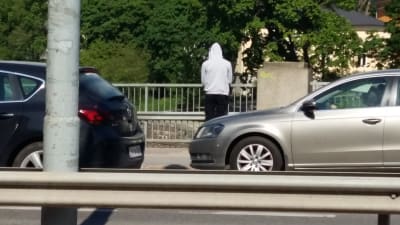Ung man i ljus munkjacka står på Domkyrkobron och blickar ut över Aura å. Två bilar susar förbi honom