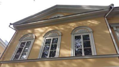 Palmetter bildar ornament ovanför tre fönster på ett gult nyklassicistiskt hus.
