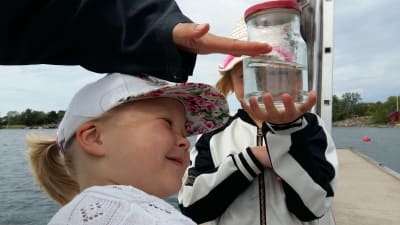 Nellie Walldén, en flicka med vit keps och vit bomullströja, tittar noga på ett vattenprov i en glasburk som hennes syster håller upp.