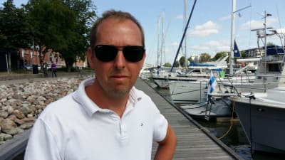 Kenneth Blomster, en man med solglasögon och vit pikéskjorta, står på en brygga med sgelebåtar.