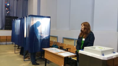 Vallokal i Ryssland. En frivillig står vid ett bord och tittar åt sidan medan man ser en man stå i ett bås och rösta.