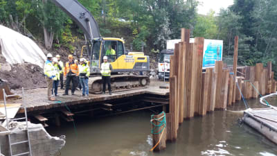 Flera personer står på en brokonstruktion på en byggarbetsplats vid vatten. I bakgrunden en grävmaskin.
