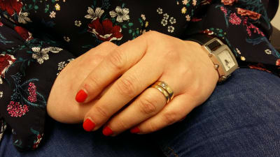 Kvinnohänder i famnen, rödlackade naglar och vigselring.