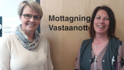 Två leende kvinnor står mot en  stägd dörr där det står "Mottagning Vastaanotto".