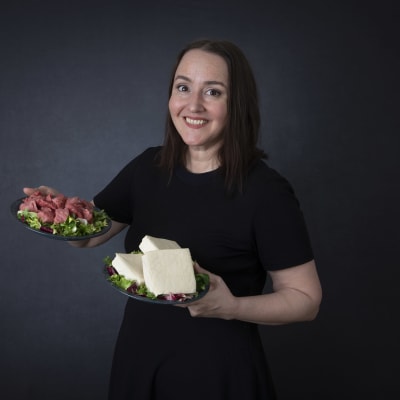 En kvinna ler glatt och serverar en tallrik med kallskurna köttprodukter och en tallrik med ost.