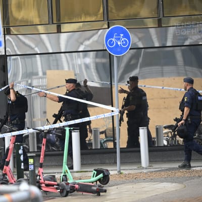 Flera tungt beväpnade poliser lyfter på ett spärrband utanför en affärsbyggnad. Intill byggnaden står flera elsparkstöttingar parkerade.