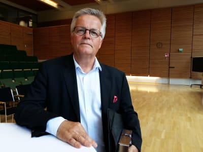 Tidigare chefredaktör Torbjörn Kevin står vid ett bord i ett auditorium med en mapp under armen.