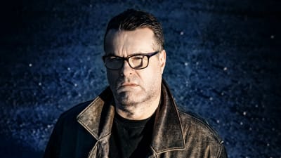 Mikael Crawfords ser allvarlig ut och står tittandes in i kameran mot en mörk bakgrund. 