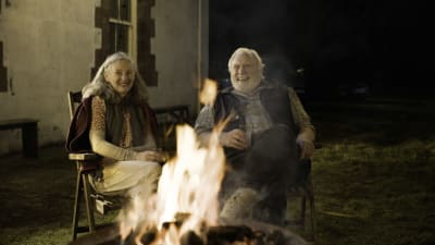 Howard (James Cosmo) och Annie (Bríd Brennan) sitter vid en brasa utomhus och ser glada ut.