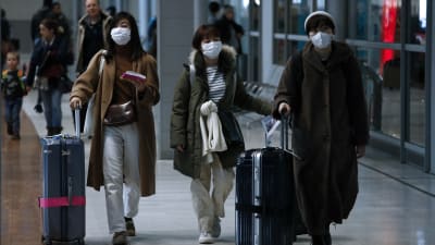 Tre damer iklädda ytterrockar och munskydd drar på var sin kappsäck i en flygplatskorridor.