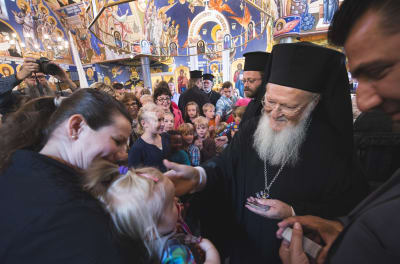 Patriark Bartholomeos I hälsar med vänlig blick på ett litet barn som är i sin mammans famn i en kyrka där både tak och väggar är målade med ikoner. Bakom patriarken syns flera andra barn och några ortodoxa präster.
