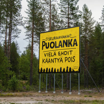 Suuri keltainen kyltti metsän laidassa, jossa lukee 'Puolanka, vielä ehdit kääntyä pois.'