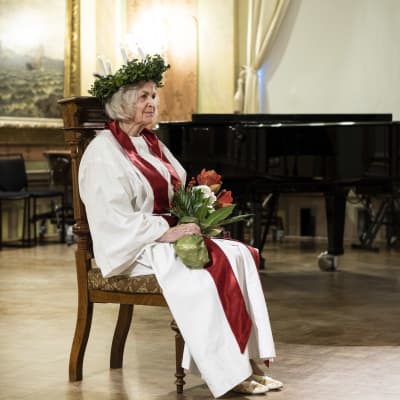 En äldre kvinna klädd som Lucia sitter på en stol