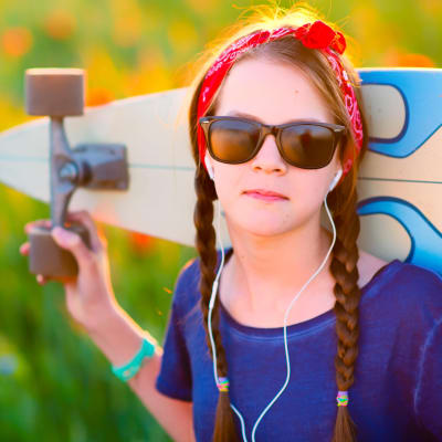 Tyttö aurinkolaseissa pitää isoa longboard-rullalautaa olkapäillään värikkään pellon laidalla.