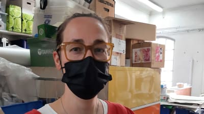 Annika Dahlsten, en dam med glasögon, svart munskydd och en röd skjorta, står framför lådor och paket i en ateljé.
