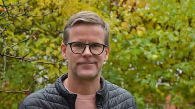 LJushårig man med kort hår och glasögon står utomhus och tittar in i kameran.