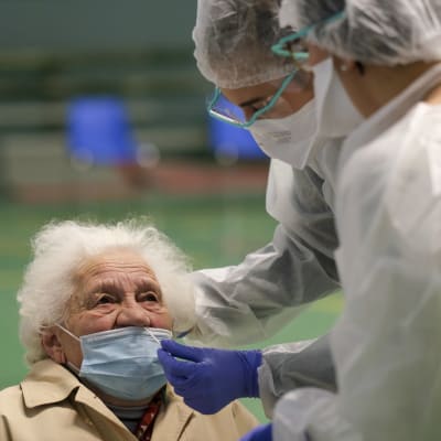 Vanha nainen saa nenäänsä testeipuikon kahdelta suojavarusteisiin pukeutuneelta henkilöltä.