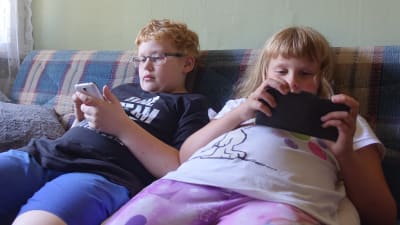 Två barn, en flicka och en pojke sitter i en rutig soffa och spelar på sina mobiltelefoner. Pojken har rött hår och glasögon och flickan har pannlugg och en vit skjorta med en bild av hunden Snoopie.