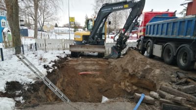En stor och djup grop mitt i en väg i centrum av Ingå. Vattenledning som gått sönder. Grävmaskin och lastbil på plats. En lekpark till vänster om vägen.