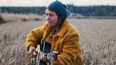 Kvinna i gul jacka och blå mössa sjunger och spelar gitarr mitt i sädesfält.