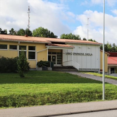 Koulurakennuksen seinällä on ruotsiksi teksti Amosparkenin koulu. Koulun edessä on lipputanko.