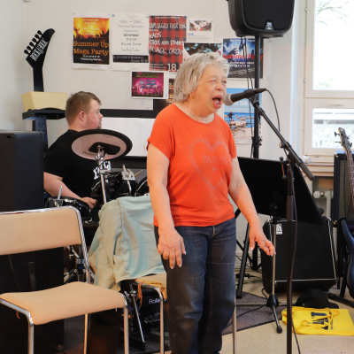 En kvinna sjunger, en man i bakgrunden spelar eltrummor och en man till höger spelar bas. 