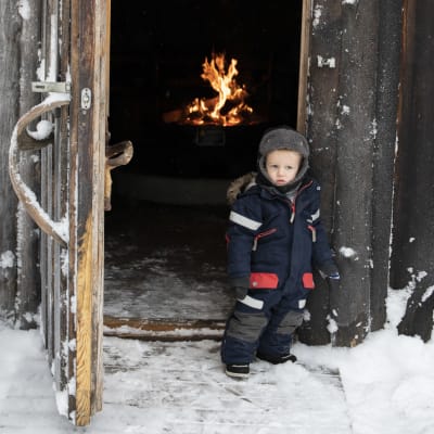En liten pojke i vinteroverall står vid dörren till en kåta.