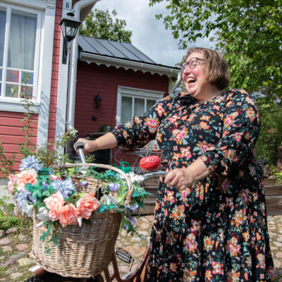 Nina Wiklund står framför det röda huset och håller i en gammalcykel med en rottingkorg fram på styret.