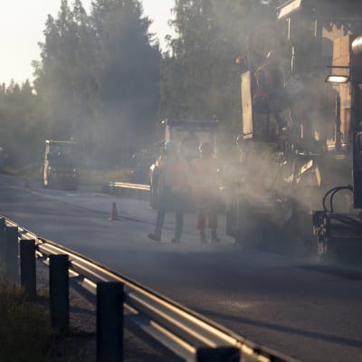 asfalttikone tekemässä asfalttia maantiellä, savun takaa erottuu asfalttityöntekijöitä kävelemässä