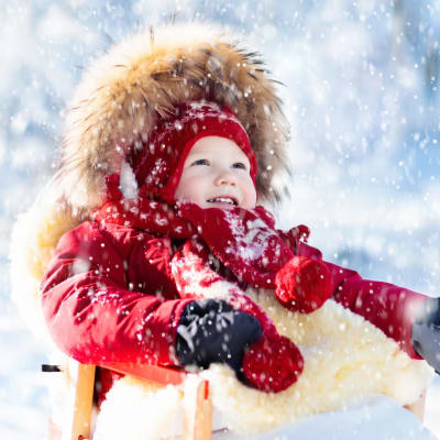 Ett barn sitter i en släde och tittar upp mot himlen medan det snöar. Solen skiner.