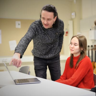 Musikläraren Tomi Norha pekar ut något på en laptop för gymnasieeleverna Oona Vahtera och Antti Lampi.