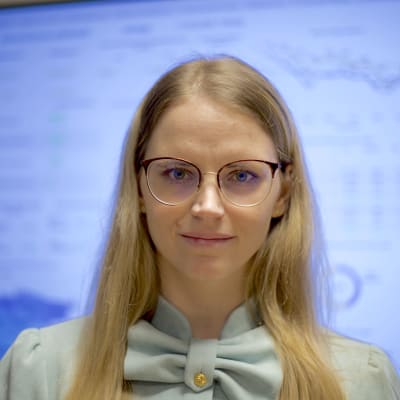 Silmälasipäinen nuori vaaleatukkainen nainen hymyilee kameraan takanaan tilastotietoa Viron koronatilanteesta.