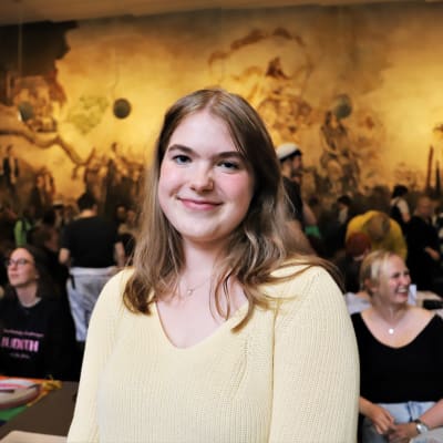 En ung kvinna med axellångt hår. I bakgrunden syns en vägg med målningar i gult och brunt föreställande studenter. 