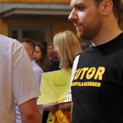 Bild av en mans t-skjorta med orden "tutor" på. Bakom står en ung kvinna och håller i en skylt med texten "Ekonomi". 